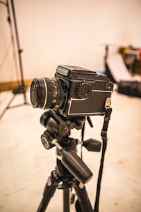 A professional camera mounted on a tripod is set up in what appears to be a photography studio. The background is slightly out of focus, with some photography equipment visible, including light stands and softboxes.