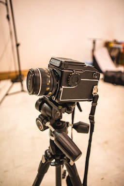 A professional camera mounted on a tripod is set up in what appears to be a photography studio. The background is slightly out of focus, with some photography equipment visible, including light stands and softboxes.