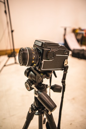 A professional camera mounted on a tripod is set up in what appears to be a photography studio. The background is slightly out of focus, with some photography equipment visible, including light stands and softboxes.