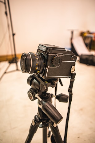 A professional camera mounted on a tripod is set up in what appears to be a photography studio. The background is slightly out of focus, with some photography equipment visible, including light stands and softboxes.