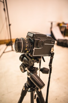 A professional camera mounted on a tripod is set up in what appears to be a photography studio. The background is slightly out of focus, with some photography equipment visible, including light stands and softboxes.