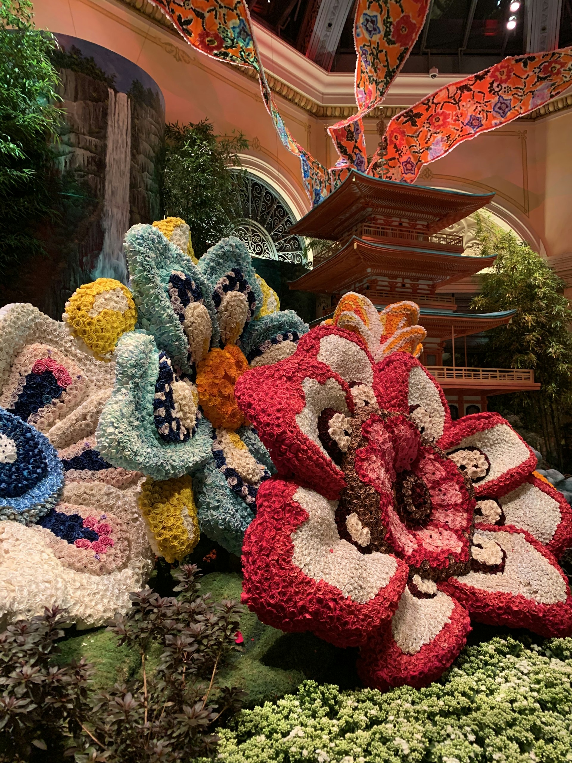The scene features a vibrant indoor garden display with large, colorful sculptures of flowers made with a multitude of small, intricately arranged materials. These flower structures display a variety of colors including red, white, blue, yellow, and orange. In the background, there is an ornate multi-tiered pagoda-style structure in a setting that resembles a greenhouse. Lush greenery and detailed decorations, including patterned cloths hung overhead, add to the rich, artistic environment.