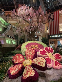 A lavish indoor display featuring a large flowering tree with cascading blossoms stands in the background. At the forefront, there are large, intricately arranged floral sculptures creating the illusion of oversized flowers. The area is well-lit with ambient lighting casting shadows over the colorful floral arrangements. A large American flag is visible in the background, next to signage for Sadelle's, indicating a possible indoor venue or garden.