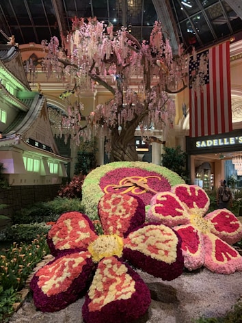 A lavish indoor display featuring a large flowering tree with cascading blossoms stands in the background. At the forefront, there are large, intricately arranged floral sculptures creating the illusion of oversized flowers. The area is well-lit with ambient lighting casting shadows over the colorful floral arrangements. A large American flag is visible in the background, next to signage for Sadelle's, indicating a possible indoor venue or garden.