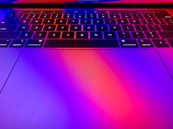 A close-up view of a laptop keyboard with vibrant lighting effects. The keys are illuminated in shades of red and blue, creating a colorful and dynamic appearance. The touchpad area also reflects this colorful lighting.
