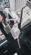 An aerial view of a cityscape featuring a prominent tower with an American flag at the top. The architecture is surrounded by modern skyscrapers and urban streets, all captured in muted colors under natural lighting.