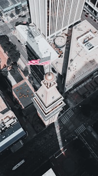 An aerial view of a cityscape featuring a prominent tower with an American flag at the top. The architecture is surrounded by modern skyscrapers and urban streets, all captured in muted colors under natural lighting.
