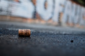 A cork lies on an asphalt road with a blurred graffiti-covered wall in the background. The photograph has a shallow depth of field, making the cork the focus, while the background is out of focus.