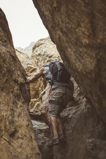 A person wearing casual climbing attire navigates through a rugged, narrow rock crevice. They are equipped with a backpack and climbing gear, including a rope looped around their shoulder. The scene captures a sense of adventure and challenge against a backdrop of textured, earthy rock formations.