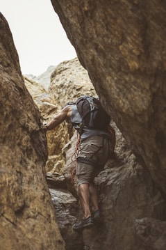 A person wearing casual climbing attire navigates through a rugged, narrow rock crevice. They are equipped with a backpack and climbing gear, including a rope looped around their shoulder. The scene captures a sense of adventure and challenge against a backdrop of textured, earthy rock formations.