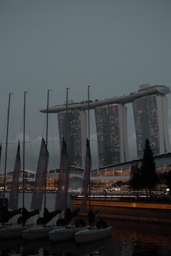A marina with several sailboats docked in the foreground, and a prominent, modern architectural structure with three towers connected at the top in the background. There is a tree and some lights illuminating parts of the scene, which appears to be taken during twilight or early evening.