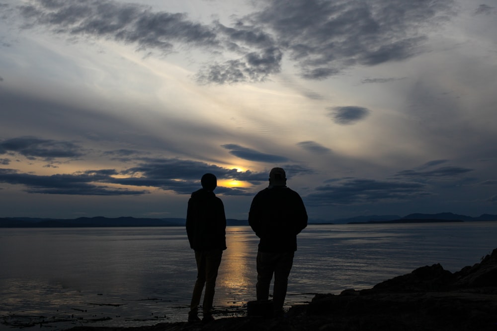 silhouette of 2 men standing on rock formation near body of water during daytime