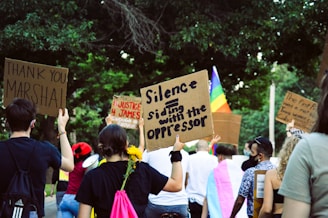 A group of people participating in a protest or demonstration, holding signs with various messages. The scene is outdoors, surrounded by trees, and includes a rainbow flag indicating a theme of equality or LGBTQ+ rights. Participants have signs addressing justice and expressing gratitude.
