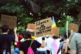 A group of people participating in a protest or demonstration, holding signs with various messages. The scene is outdoors, surrounded by trees, and includes a rainbow flag indicating a theme of equality or LGBTQ+ rights. Participants have signs addressing justice and expressing gratitude.