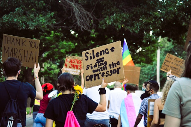 A group of people participating in a protest or demonstration, holding signs with various messages. The scene is outdoors, surrounded by trees, and includes a rainbow flag indicating a theme of equality or LGBTQ+ rights. Participants have signs addressing justice and expressing gratitude.
