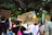A group of people participating in a protest or demonstration, holding signs with various messages. The scene is outdoors, surrounded by trees, and includes a rainbow flag indicating a theme of equality or LGBTQ+ rights. Participants have signs addressing justice and expressing gratitude.