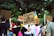 A group of people participating in a protest or demonstration, holding signs with various messages. The scene is outdoors, surrounded by trees, and includes a rainbow flag indicating a theme of equality or LGBTQ+ rights. Participants have signs addressing justice and expressing gratitude.