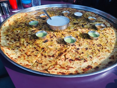 A large circular bread, possibly a paratha or pancake, served on a huge metal tray with small bowls of various sauces or dips arranged around its center. The bread appears to be topped with herbs and spices.