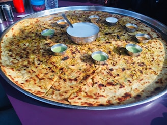 A large circular bread, possibly a paratha or pancake, served on a huge metal tray with small bowls of various sauces or dips arranged around its center. The bread appears to be topped with herbs and spices.