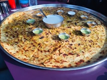 A large circular bread, possibly a paratha or pancake, served on a huge metal tray with small bowls of various sauces or dips arranged around its center. The bread appears to be topped with herbs and spices.