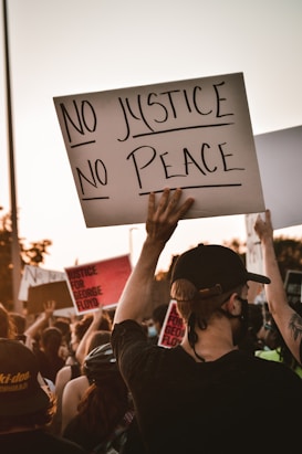A crowd of people is participating in a protest. One prominent sign held up by a person in the foreground reads 'No Justice No Peace'. Other signs referencing justice and George Floyd are visible in the background. The crowd appears diverse, and many individuals are wearing face masks.