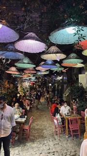 A lively outdoor dining setting with colorful woven lampshades hanging overhead. People are seated at wooden tables along a narrow, cobblestone street, creating a cozy and vibrant atmosphere. Lush greenery and flowers adorn the surrounding area, enhancing the charming ambiance.