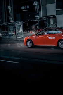 A brightly colored orange taxi with 'Beck' written on its side is captured driving through a dimly lit urban street at night. The scene includes a modern bus stop with illuminated advertisements and the reflection of city lights in tall building windows.