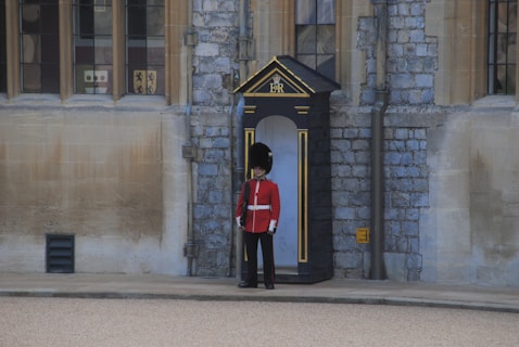 A traditional guard wearing a red uniform and black bearskin hat stands in a black and gold sentry box. The background features a historic stone building with large glass windows.