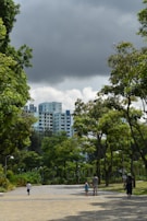 A pathway leading through a green park with lush trees on both sides. In the background, tall residential buildings are visible under a cloudy sky. Several people casually walk along the path, including a child wearing blue and a couple of adults.
