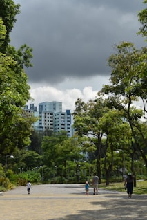 A pathway leading through a green park with lush trees on both sides. In the background, tall residential buildings are visible under a cloudy sky. Several people casually walk along the path, including a child wearing blue and a couple of adults.