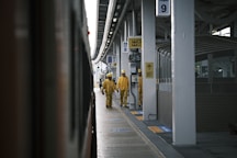 Two workers in yellow uniforms and helmets walk along a train platform. The platform is covered, with multiple signs and a train visible on the left. The environment appears to be industrial, with a focus on public transit.