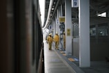 Two workers in yellow uniforms and helmets walk along a train platform. The platform is covered, with multiple signs and a train visible on the left. The environment appears to be industrial, with a focus on public transit.