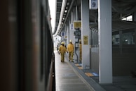 Two workers in yellow uniforms and helmets walk along a train platform. The platform is covered, with multiple signs and a train visible on the left. The environment appears to be industrial, with a focus on public transit.