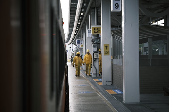 Two workers in yellow uniforms and helmets walk along a train platform. The platform is covered, with multiple signs and a train visible on the left. The environment appears to be industrial, with a focus on public transit.