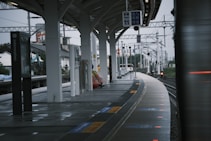 A modern train station platform with several pillars supporting a curved roof. The platform features information signs and electronic boards displaying train schedules. Sparse seating and a few people are visible in the distance, with a cityscape in the background. The tracks curve gently away, and the atmosphere appears overcast.