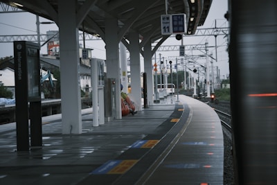 A modern train station platform with several pillars supporting a curved roof. The platform features information signs and electronic boards displaying train schedules. Sparse seating and a few people are visible in the distance, with a cityscape in the background. The tracks curve gently away, and the atmosphere appears overcast.