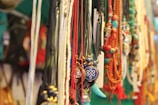 A collection of colorful necklaces with various beads and pendants is displayed. The strings are made of different materials and colors, including blue, red, and green. Notable pendants include a blue and gold circular design and a turquoise tusk-like charm. The background is blurred, highlighting the vibrant jewelry.