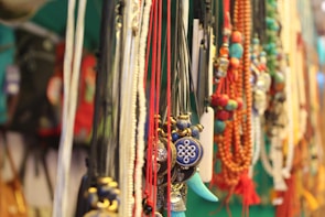 A collection of colorful necklaces with various beads and pendants is displayed. The strings are made of different materials and colors, including blue, red, and green. Notable pendants include a blue and gold circular design and a turquoise tusk-like charm. The background is blurred, highlighting the vibrant jewelry.