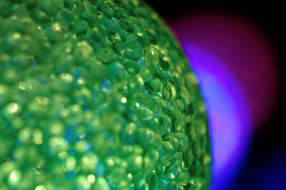 A close-up macro photograph featuring numerous small green beads or crystals that create a textured surface. The background is blurred, showcasing a gradient of dark colors with a noticeable purple glow.