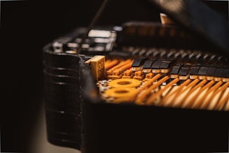 A close-up view of the internal mechanics of a piano, focusing on the strings and hammers. The image reveals intricate details of the instrument with brass-colored strings and black felted hammers. The lighting highlights the textures and craftsmanship.