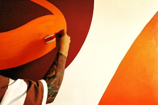 A person with a tattooed arm is painting a wall using a paint roller. The wall features curved stripes in shades of orange and red, with the background being predominantly white.