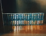 A neatly arranged collection of books with identical dark blue covers and gold lettering sits on a wooden shelf. Each book is uniformly positioned, creating a visually appealing display.