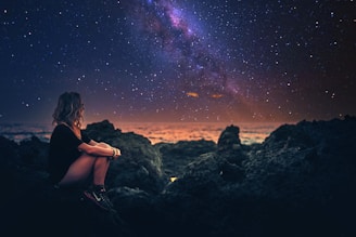 A woman sits on rocky terrain by the ocean, gazing at a vibrant night sky filled with stars and the Milky Way. The horizon glows with a dim, warm light, creating a serene and contemplative atmosphere.
