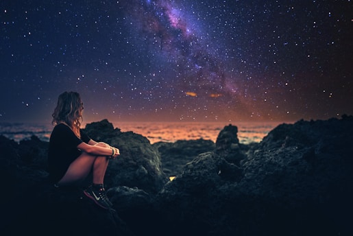 A woman sits on rocky terrain by the ocean, gazing at a vibrant night sky filled with stars and the Milky Way. The horizon glows with a dim, warm light, creating a serene and contemplative atmosphere.