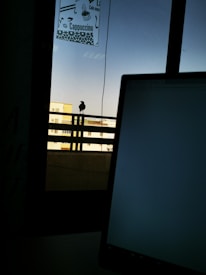 A silhouetted bird perched on a metal railing against a clear sky. In the foreground, a partial view of a laptop screen is visible, with a window frame and a sign displaying a coffee menu above. Buildings can be seen in the background.