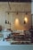 A cozy coffee shop interior with a wooden menu board on the wall, displaying options for specialty coffee, loose leaf tea, and hot chocolate. The counter has a glass display case filled with assorted pastries, and there are various coffee-making supplies and packaged goods arranged neatly. Overhead pendant lights hang, adding a warm atmosphere.