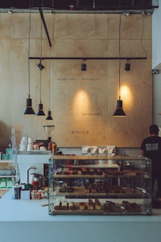 A cozy coffee shop interior with a wooden menu board on the wall, displaying options for specialty coffee, loose leaf tea, and hot chocolate. The counter has a glass display case filled with assorted pastries, and there are various coffee-making supplies and packaged goods arranged neatly. Overhead pendant lights hang, adding a warm atmosphere.