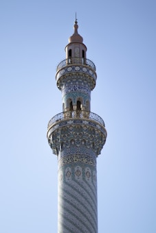 A tall, intricately decorated minaret with ornate Islamic patterns and calligraphy. The structure features blue and green tiles with geometric and floral designs, capped by a golden dome with a finial. The sky in the background is clear and blue.