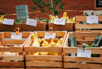 A farmer's market display featuring a variety of vegetables in wooden crates. There are signs indicating the types and prices, such as shishito peppers, zucchini, candy onions, and summer squash. The background consists of a red brick wall and green foliage.