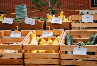 A farmer's market display featuring a variety of vegetables in wooden crates. There are signs indicating the types and prices, such as shishito peppers, zucchini, candy onions, and summer squash. The background consists of a red brick wall and green foliage.